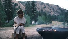 a woman sitting next to a campfire writing on a piece of paper in her hand