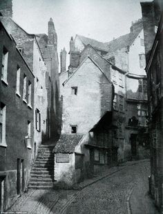 black and white photograph of an old street with buildings on both sides, looking up at the sky