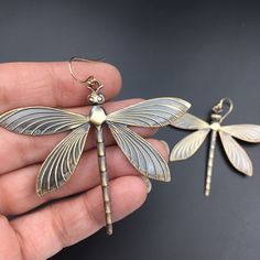 a person holding two dragonfly shaped earrings in their hand, one is gold and the other is silver