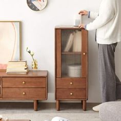 a woman standing next to a wooden cabinet in a living room with a clock on the wall