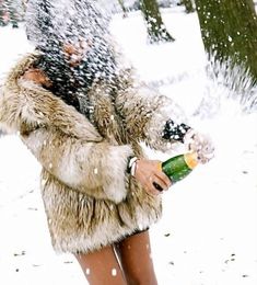 a woman in a fur coat holding a bottle and spraying snow on her face while standing next to a tree