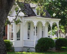 a small white house with a porch on the front and side of it, surrounded by trees