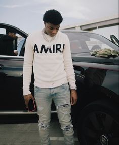 a man standing in front of a car wearing ripped jeans and a sweater with the word amir on it