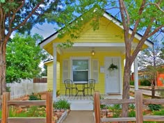 a small yellow house with two chairs on the front porch and table in the yard