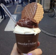 a person holding up a chocolate gelato with waffles on top in the street