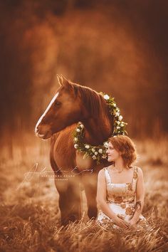 a woman sitting in the grass next to a horse with a wreath on it's head