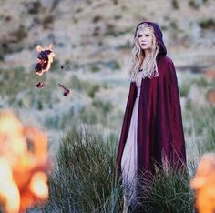 a woman in a red cape standing in tall grass with fire coming out of her mouth