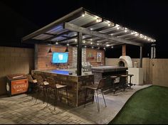 an outdoor kitchen and grill area at night with lights on the roof, bar stools and television