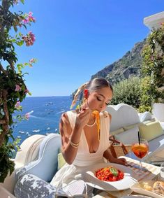 a woman sitting at an outdoor table with food and drinks in front of the ocean