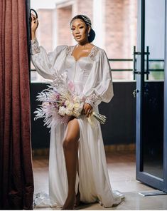 a woman standing next to a window wearing a white dress and holding a flower bouquet