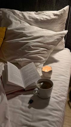 a cup of coffee and an open book on a bed with white linens, next to a lit candle