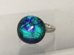 This dichroic fused glass stone set on an adjustable sterling silver 925 ring band (hallmarked). The dichroic glass is a deep emerald green dichroic glass stone which has an abstract deep blue/purple ripple pattern running through it. The glass also changes from bright emerald green to blue/green as it moves in the light or is seen from an angle giving added interest. The stone is round in shape measuring approximately 12 mm in diameter.  Unadjusted, the ring shank measures UK M (US 6.5) and can Iridescent Cabochon Ring As Gift, Iridescent Cabochon Rings As Gift, Modern Iridescent Round Rings, Modern Iridescent Round Ring, Adjustable Iridescent Rings, Ripple Pattern, Green To Blue, Cerise Pink, Ring Shank