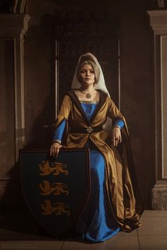 a woman dressed in medieval clothing sitting on a chair holding a shield and wearing a tiara