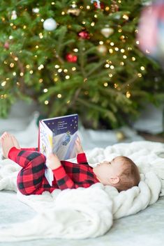 a baby is laying on a blanket reading a book in front of a christmas tree