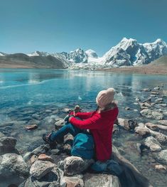 a person sitting on some rocks by the water with mountains in the backgroud