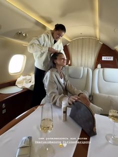 a woman sitting at a table in an airplane with wine glasses on it and another person standing next to her