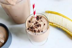 a smoothie in a glass next to a banana on a white table with two straws