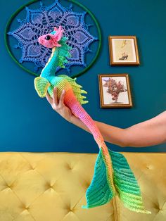 a woman holding a crocheted seahorse in her hand with pictures on the wall behind her