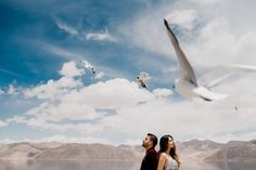 a man and woman looking at seagulls flying in the sky over water with mountains in the background