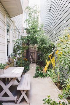 a wooden table sitting next to a white house with lots of plants on the side