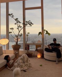 two people sitting on couches in a living room with large windows overlooking the city