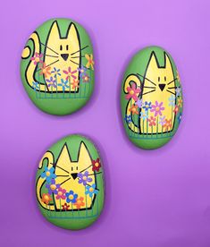 three painted rocks with cats on them sitting in front of a purple background and one has flowers