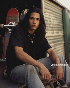 a man with long hair sitting on the ground next to his skateboard and wearing jeans