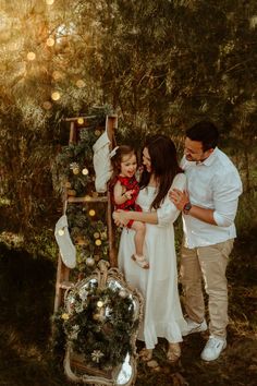 a man and woman holding a baby in front of a ladder with christmas decorations on it