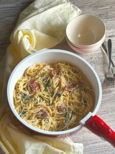 a bowl of pasta with spinach and tomato sauce on a wooden table next to a red spoon