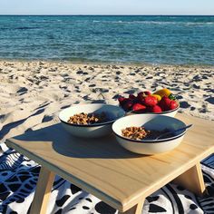 two bowls of cereal and strawberries sit on a table at the beach near the ocean