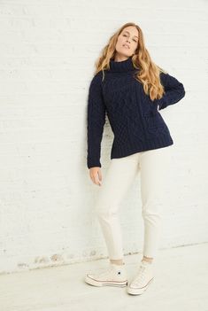 a woman leaning against a wall wearing white pants and a blue cable knit turtle neck sweater