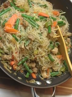 a wok filled with noodles and vegetables on top of a wooden cutting board next to a spatula