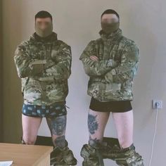 two men in camouflage jackets and shorts standing next to each other with their arms crossed