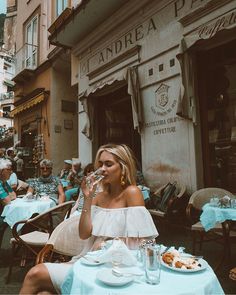 a woman sitting at a table in front of a building drinking from a water bottle