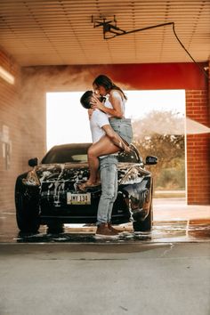 a man and woman standing on the hood of a black car in front of a garage