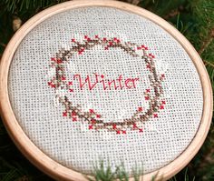 a cross stitch ornament with the word winter written in red and surrounded by pine needles