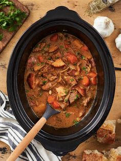 a stew in a crock pot on a wooden table with garlic bread and parsley