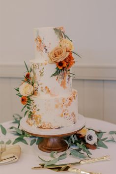 a wedding cake with flowers and greenery sits on a table next to silverware