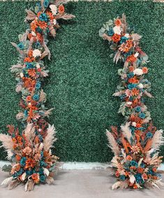 two tall floral arrangements with orange, blue and green flowers on the side of a wall