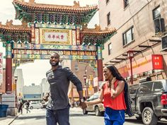 a man and woman holding hands walking under an archway on a city street with cars in the background