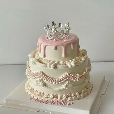 a three tiered cake with pink icing and a crown on top is sitting on a white box