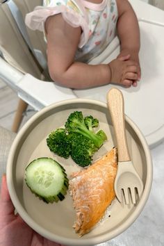 a baby sitting in a highchair holding a plate with fish, broccoli and cucumbers