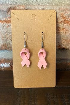 the pink ribbon earrings are on display in front of a brick wall and wooden floor