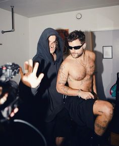 a man with no shirt and sunglasses sitting next to another man in a black hoodie