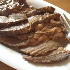 a white plate topped with sliced beef and onions next to a fork on a wooden table