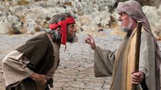 two men with red bandanas talking to each other on the street in front of some rocks