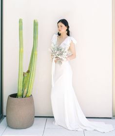 a woman standing next to a tall cactus in a white dress and holding a bouquet