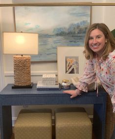 a woman standing next to a blue table with two stools and a painting on the wall behind her