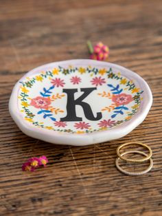 two gold wedding rings sitting on top of a wooden table next to a plate with the letter k