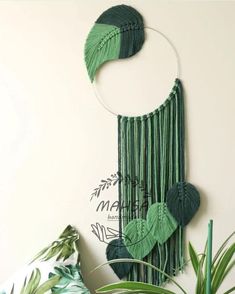 a wall hanging made with green yarn and leaves on a white wall next to a potted plant